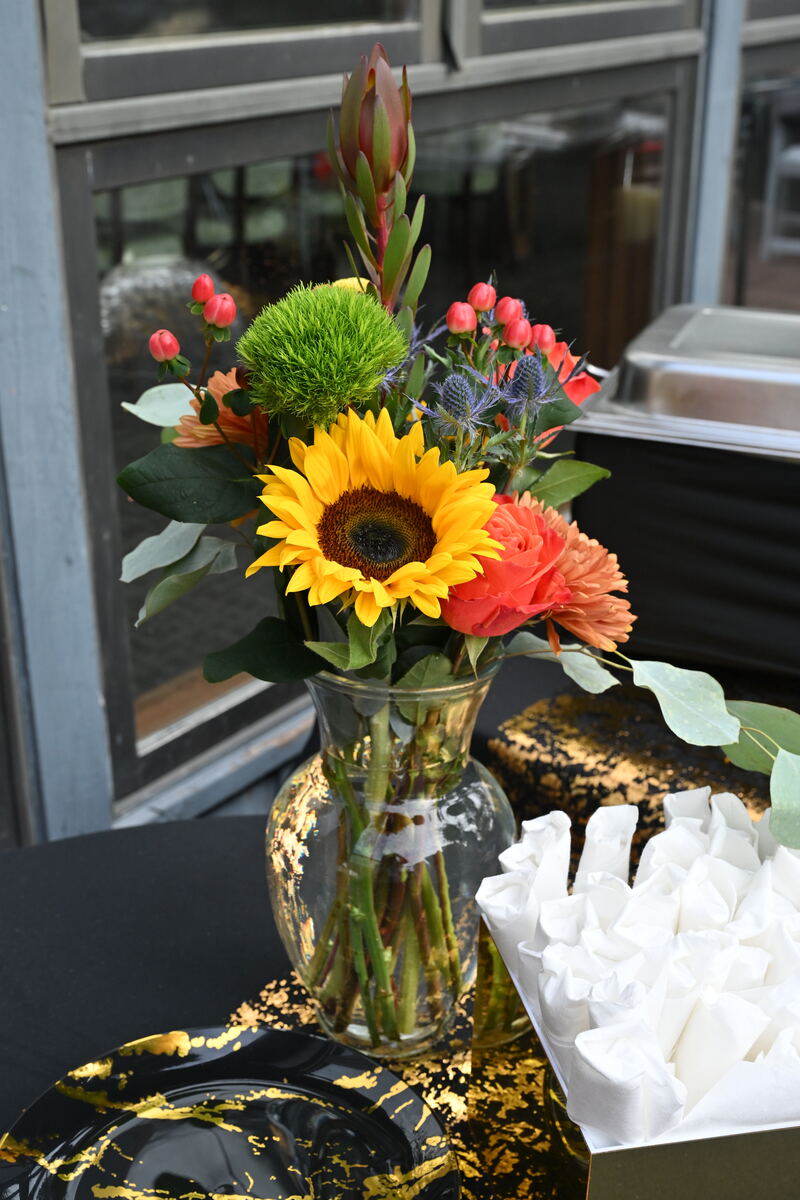 A tall arrangement of sunflowers, orange roses and greenery in a glass vase on a gold-draped table