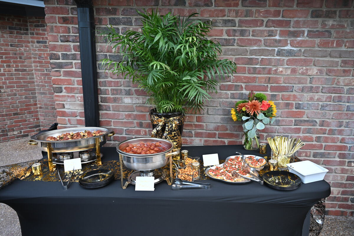 A buffet table against the brick wall, chafing dishes and platters laid out beneath a palm and a vase of flowers